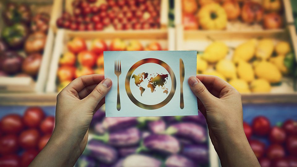 Global food issues. Hands holding a paper sheet with world map in a plate with knife and fork icon over market shelves background.